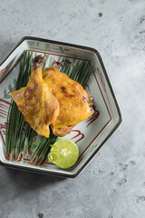 A piece of grilled chicken rests on pine needles in a hexagonal bowl with a lime half. The gray stone background and clean styling suggest refined, minimalist plating.