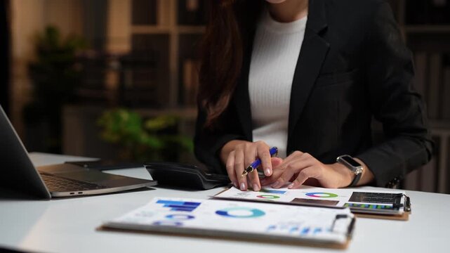 Focused accountant working overtime in a modern office, using a calculator to check financial data on printed charts and graphs, analyzing company performance and making business calculations