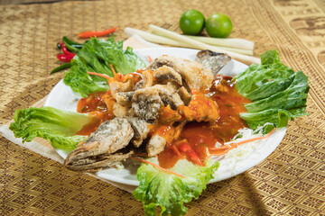 A deep fried whole fish is served with a glossy tangy chili sauce and lettuce on a patterned table. Limes, chilies and lemongrass in the background suggest bold tropical flavors.