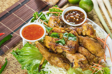 A platter of grilled chicken pieces rests on shredded greens with two dipping sauces. Cilantro and chili add freshness, styled on patterned cloth beside bamboo mats.