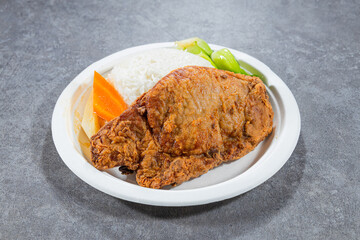 A golden fried pork chop sits with white rice, carrot and greens on a round disposable plate. The simple presentation suggests a casual and comforting set meal.