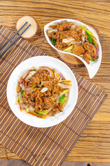 Two plates of stir fried beef are topped with onion and scallion. Set over bamboo mats on a wooden table, the glossy slices suggest a savory, homestyle dish served for sharing.