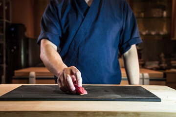 At the counter, a chef presents a fresh nigiri over a dark board. The scene emphasizes neat service, hygiene and attention to detail in traditional sushi making.