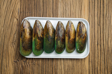 Six green mussels are arranged in a white tray on rustic wood. The shells show natural brown and teal tones, presenting a clean seafood ingredient ready for cooking.