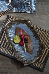 On a textured ceramic plate, a leaf wrapped piece of grilled fish is paired with citrus and a small garnish stick. Woven mats and patterned cloth frame a refined, Japanese inspired plating.