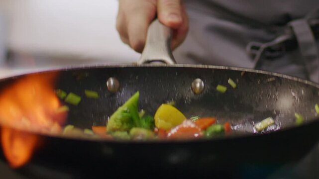 Close-up view of restaurant chef tossing stir fry vegetables in hot skillet as flames burning during flambe cooking process. Slow motion