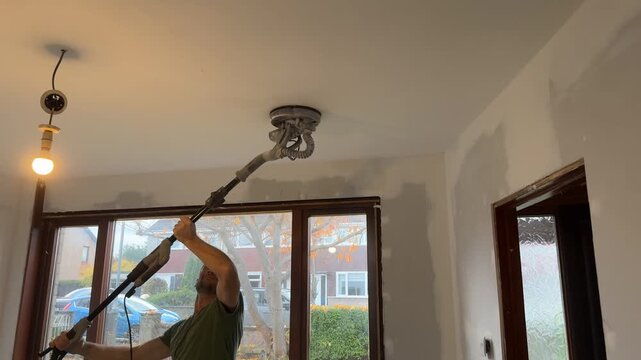 A European man sands a ceiling with a sander. Ceiling repair work. A sander in action, medium shot. A construction worker sands a ceiling. Home renovation and restoration.