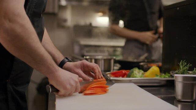 Chef hands slicing carrot on prep board in restaurant kitchen while his coworker flambeing food in skillet in background. Close-up shot