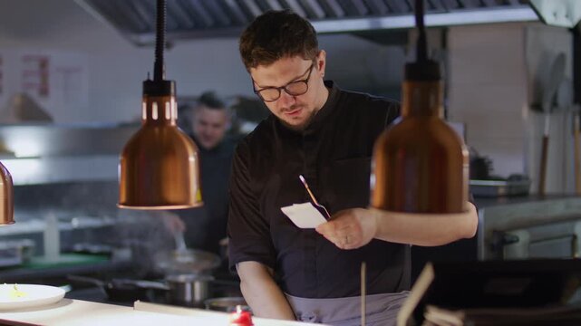 Chef reading order ticket, giving tasks to his team and taking notes while coordinating food preparation in busy restaurant kitchen, his colleague flambeing with high flame in background