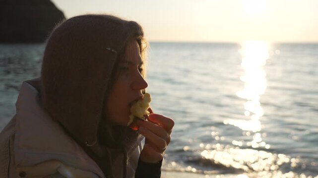 Woman eating apple core near the sparkling sea waves at sunset, enjoying healthy snack outdoors.