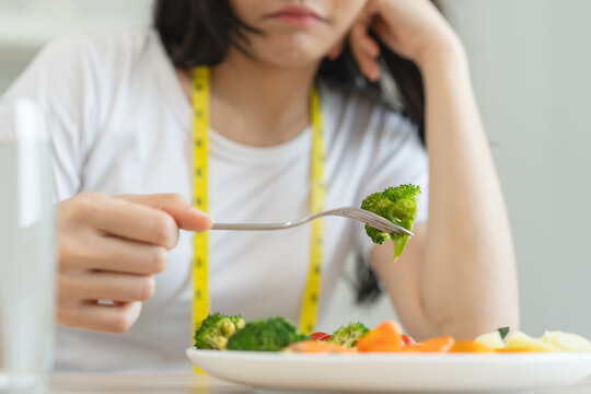 unhappy asian women is on dieting time looking at broccoli on the fork. girl do not want to eat vegetables and dislike taste of broccoli.