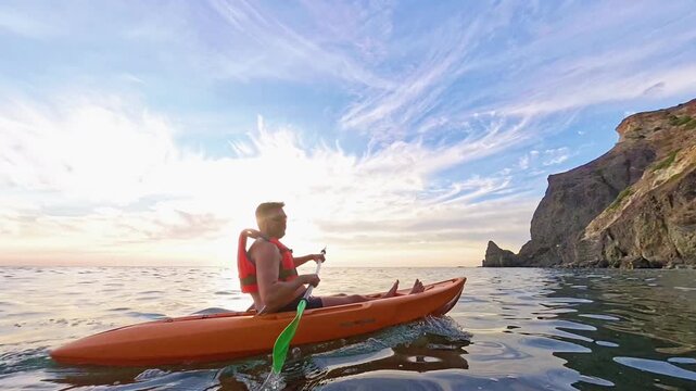 Kayak sunset ocean featuring a man paddling an orange boat on calm water near a rugged cliff coastline at golden hour