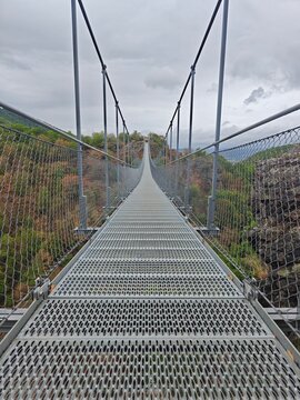 Long modern metal suspension bridge with a unique perforated walkway extends across a scenic valley.