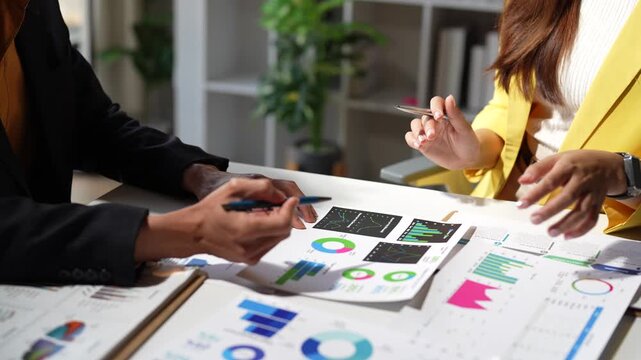 Two business professionals sitting at a desk, discussing and analyzing financial reports with various charts and graphs, planning a new marketing strategy and pointing to key data points