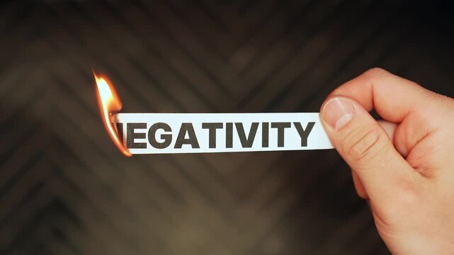 Close-up of a hand holding a paper strip labeled &ldquo;NEGATIVITY&rdquo; as the edge burns with flame on a dark background. Letting go of toxic thoughts concept with dramatic copy space.
