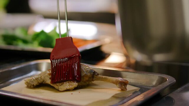 Close-up view of unrecognizable chef brushing sauce onto roasted duck leg with silicone brush on baking tray while cooking gourmet dish in restaurant kitchen