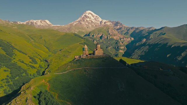 Drone view of ancient Gergeti Trinity Church sits atop a green hill, with majestic snow-capped Mount Kazbek (Kazbegi) rising in the background near Stepantsminda, Georgia