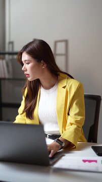 Confident Asian businesswoman in a yellow suit jacket sitting at her desk, typing on a laptop computer, and analyzing financial data charts in a modern corporate office workplace