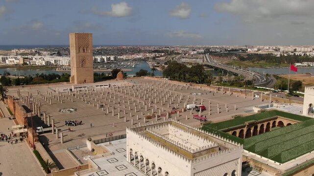 A professional HD high-angle aerial view of the Hassan Tower square in Rabat, Morocco. The footage showcases the geometric rows of ancient pillars and the iconic minaret, with the Bou Regreg river and