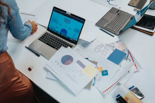 A business team member works at a white desk with a laptop displaying a flowchart and printed reports. The desk is scattered with charts, markers, and devices in a bright office.