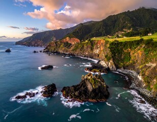 Fototapeta premium Aerial shot of coastal scenery, with rugged cliffs, dark rocks in the water, and green forested mountains, under a colorful sky