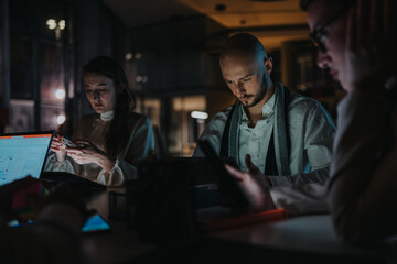 Group of professionals work late at a dimly lit office, using laptops and tablets. The scene shows focus and collaboration around a desk with screens. © qunica.com