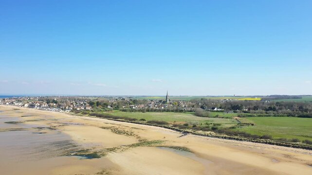 Wide aerial shot of Juno Beach and the village of Bernieres sur Mer, Normandy, France, with sandy shoreline, green fields, and a clear blue sky.