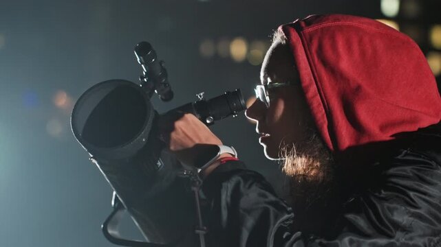 night sky stargazer with telescope, red hood and glasses adjusting eyepiece against city bokeh, focused amateur astronomer studying star fields, tripod mount and silhouette profile, cold evening