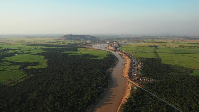 Drone shot of the river and lush landscape surrounding the floating village of Chong Kneas near Siem Reap, Cambodia. Traditional houses line the waterway amid green fields and distant hills.