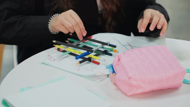 asian student arranging colorful markers on round table, pink pouch and notebook beside hands, closeup sorting pens and highlighters, focused methodical mood, soft indoor light and cozy studybreak