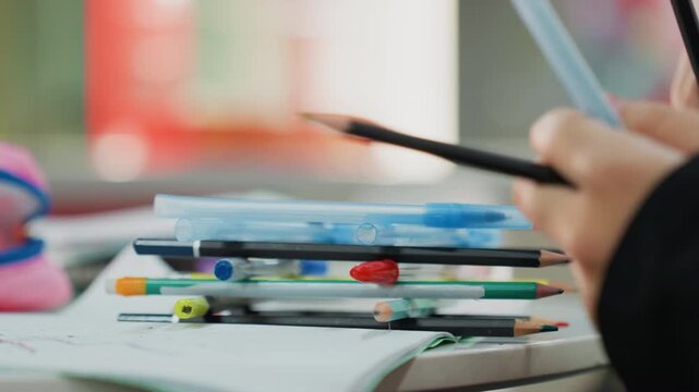 student hands arranging colorful pencils pens and notebook stacked on desk, closeup of grip and fingertip movement, soft classroom bokeh, casual study mood, student focused on organization