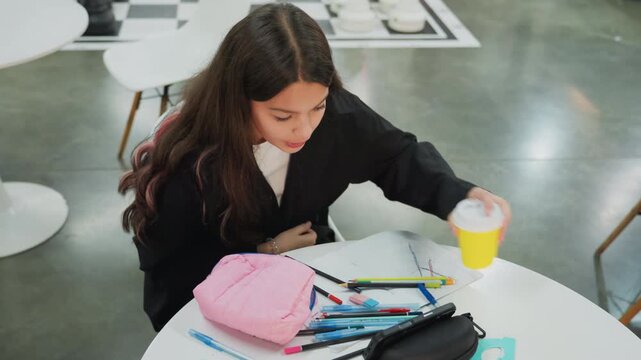teen girl studying at cafe table, surrounded by pink pencil pouch, scattered pens and papers and yellow takeaway cup, focused expression as she reads and sips during homework session in modern