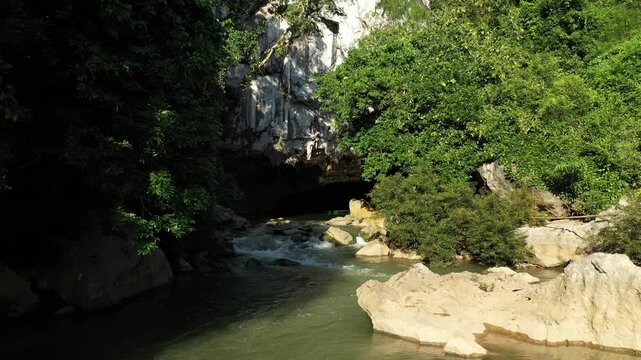 Scenic river enters the limestone entrance of Kong Lor Cave surrounded by lush green vegetation and rocky cliffs in Laos.