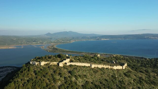 Aerial view of the historic Navarino Castle ruins atop a green hill with sweeping views of the bay and distant mountains in Greece. Clear blue sky and tranquil water create a scenic landscape.