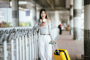 Obraz premium Young Asian woman traveler standing at an airport terminal with a yellow suitcase, holding coffee and smartphone, wearing headphones while waiting for departure.