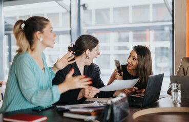 Three businesswomen collaborate and react excitedly while reviewing documents at a meeting table. Colleagues discuss ideas and celebrate progress during a lively office collaboration.