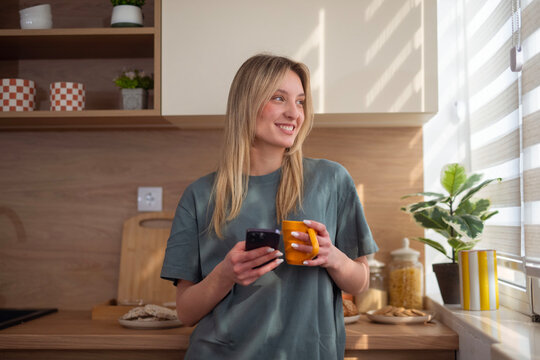 Woman smiling holding smartphone and coffee mug in kitchen