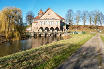 Obraz premium The massive pumping station rises beside the calm canal while fresh green tones and a willow tree suggest early spring. Architecture and hydraulic engineering in clear perspective.