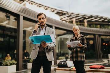 Fototapeta premium Two professionals outside a modern building review documents and speak on the phone, conveying collaboration and business activity.
