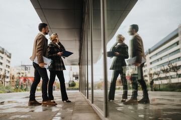 Two professionals stand outside a glass building, holding folders and a notebook. Their reflection appears on the glass as they discuss in an urban, business setting.