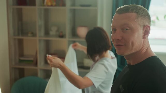 man receiving microcurrent facial massage, therapist prepares towel and electrode in cozy treatment room with soft lamp light, curtained window, decorative shelf and ceramic pots client smiles calmly