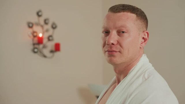 white man in robe by sconce, spa client smiles between profile and closeup during professional microcurrent facial, warm candlelight on beige wall, modern lamp and mirror in background, relaxed