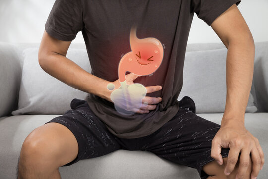 Photo of a man sitting on a sofa holding his abdomen with a cartoon stomach graphic illustrating acid reflux, bloating, and gastric discomfort at home.