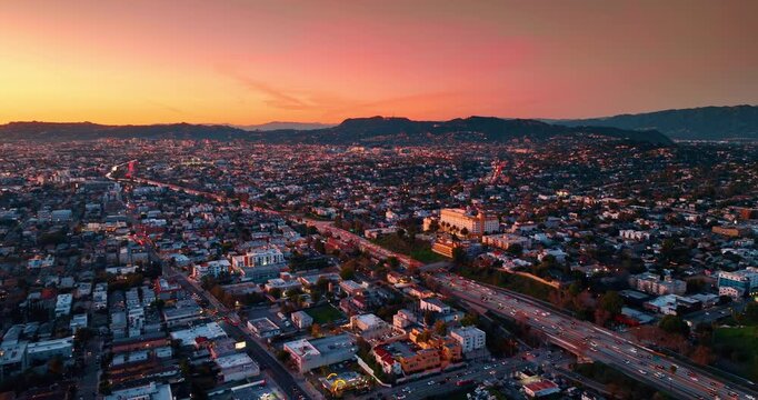 Approaching multi-lane highway with hectic traffic crossing the vast city scenery. Panorama of Los Angeles, California, USA against pink sky. Mountain silhouettes along the horizon. Aerial view.