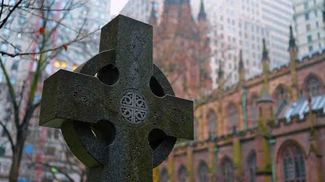 Celtic cross headstone in Trinity Church graveyard New York. Close up of a stone cross memorial in an urban cemetery with tall buildings in the background.
