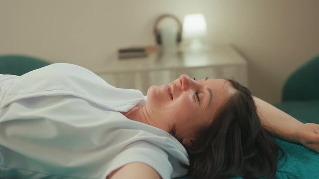 caucasian woman lying on treatment bed, receiving gentle microcurrent facial with eyes closed and relaxed smile, teal blanket and bedside lamp create soft ambient glow, clinical spa setting