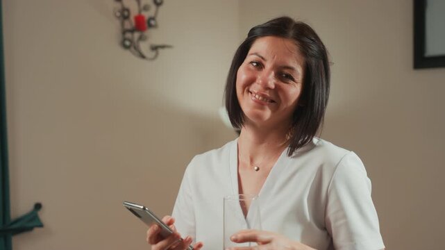 caucasian woman therapist holding microcurrent device, smiling softly in relaxed home spa setting wearing white uniform, preparing facial massage treatment with glass of water on side, warm lamp