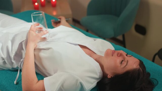 Caucasian woman reclining on spa table holding glass and remote, candles glowing in background, white robe, warm ambient lighting, microcurrent facial device nearby, relaxed smile, luxury pampering