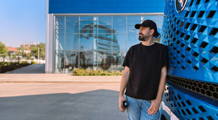 A young man is a professional driver near a blue truck. A handsome guy, a transportation worker stands next to a large truck car © Anna