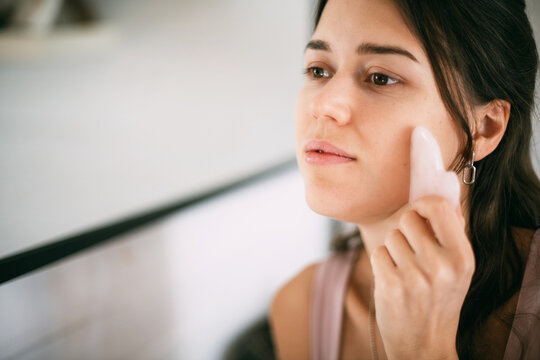 A young woman makes a facial massage with a gouache scraper at the mirror at home.